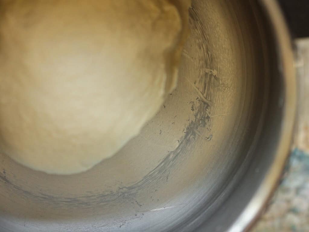 A close-up of dough in a mixing bowl. The dough is smooth and pale in color, partially stuck to the side of the stainless steel bowl, suggesting its being prepared for baking.